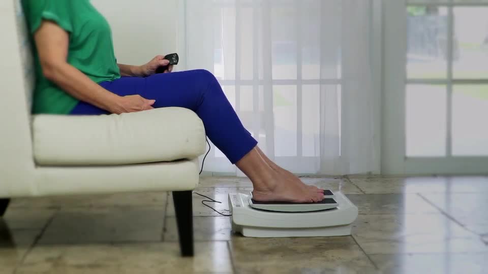 A woman in blue leggings and a green top sits on a white armchair, holding a remote. Her bare feet rest on a white platform that appears to be vibrating.