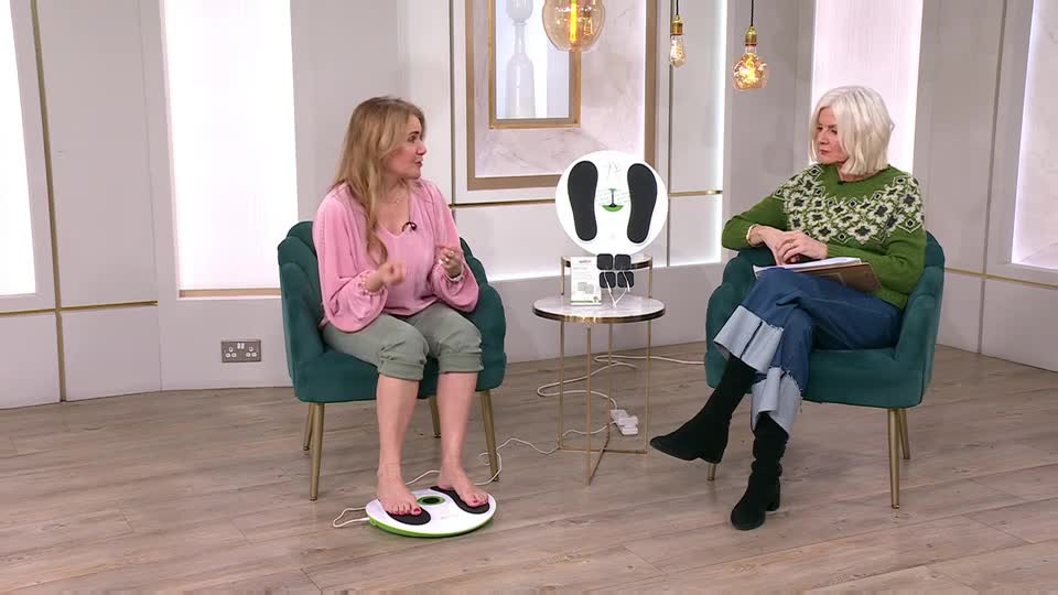 A woman in a pink top is demonstrating a foot massager on a QVC Beauty set. Another woman in a green sweater watches her, holding a tablet.
