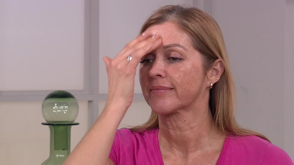 Her hand glides across her forehead, smoothing something onto her skin. The QVC presenter is demonstrating a beauty product, her expression focused.