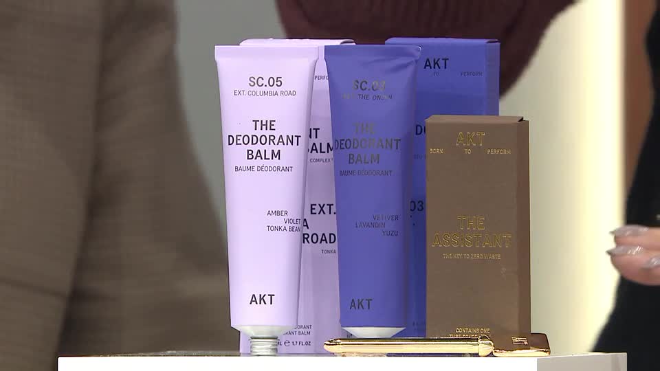 Two tubes of deodorant balm, one lilac and one lavender, stand next to a brown box labelled "The Assistant." A gold applicator rests in front of them.