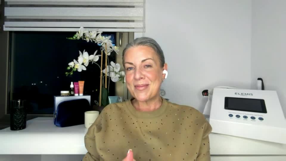 A woman with grey hair, wearing a tan sweatshirt, smiles directly at me. Beside her, a white Elemis device sits on a shelf, its screen displaying a dark interface.