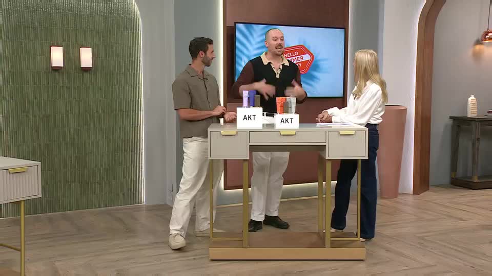 A man in a patterned shirt gestures animatedly while standing behind a desk with QVC Beauty products. A woman in a white blouse and dark trousers stands opposite him, listening intently.