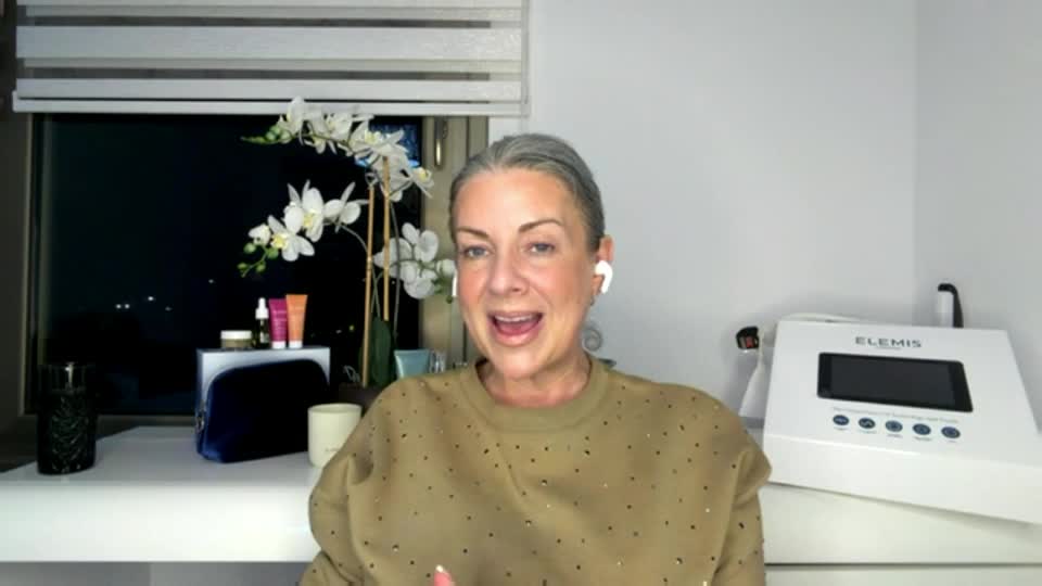 A woman with grey hair and white earbuds is speaking animatedly. Behind her, a window shows a dark night sky with distant lights, and a white Elemis machine sits on a shelf.