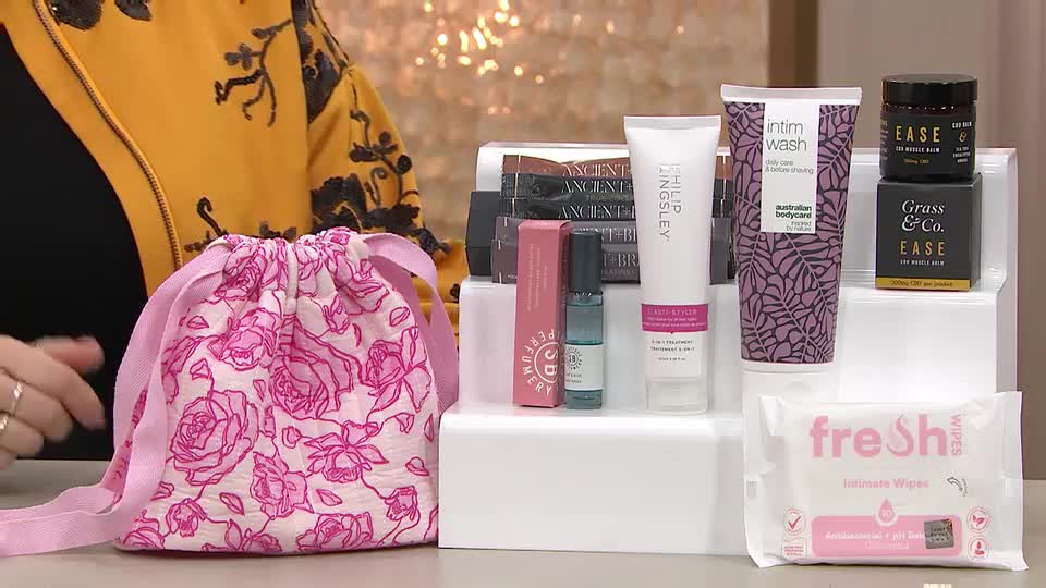 A hand reaches for a floral drawstring bag. Behind it, a collection of beauty products is neatly arranged on white risers.