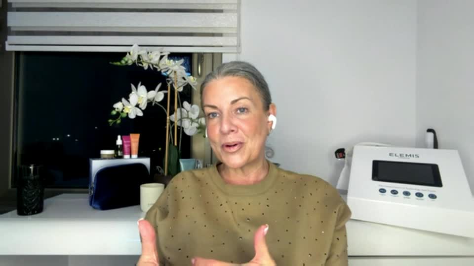 A woman with grey hair and a tan sweatshirt is speaking, her hands gesturing animatedly. Behind her, a window shows a dark night sky with distant lights, and a white Elemis machine sits on a shelf.