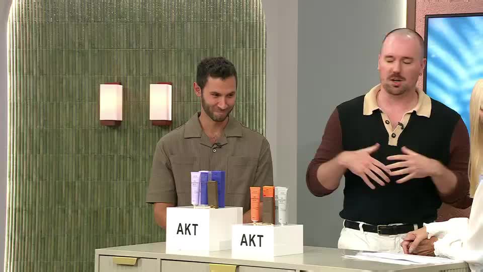 A man in a brown shirt smiles as he stands next to a display of AKT beauty products. To his right, another man gestures with his hands while talking, also with products on display.