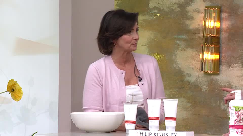 A woman in a pink cardigan stands beside a white bowl, looking towards a display of Philip Kingsley products. Her hand gestures towards a large pump bottle on the right.