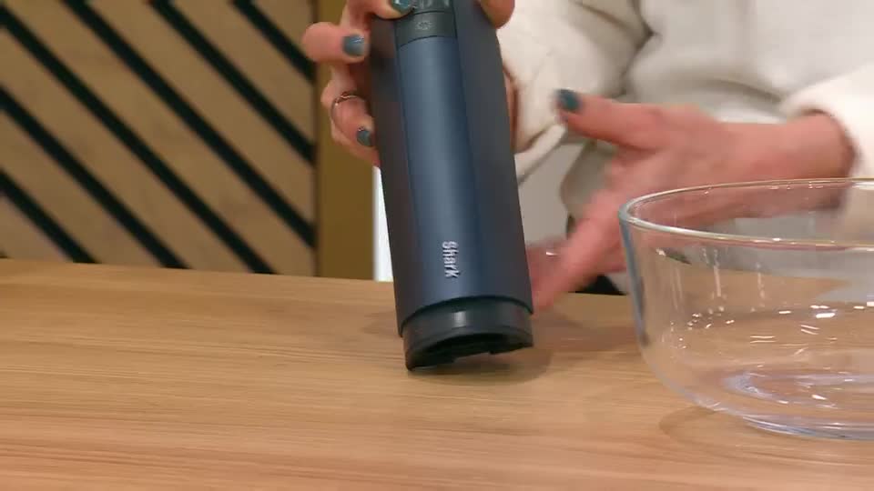 A dark blue Shark vacuum cleaner is being held over a glass bowl. The presenter's hands, with teal nail polish, guide the device. A dark blue Shark vacuum cleaner is being held over a glass bowl. The presenter's hands, with teal nail polish, guide the device.