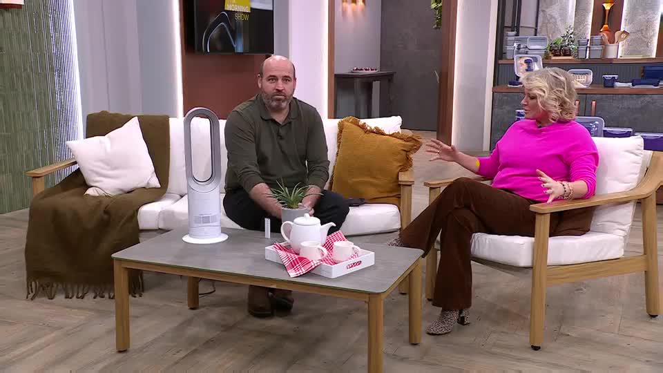 A man sits on a white sofa next to a tall, white fan, while a woman in a bright pink top gestures animatedly from a wooden armchair. A small tea set rests on the coffee table between them, looking ready for a break. A man sits on a white sofa next to a tall, white fan, while a woman in a bright pink top gestures animatedly from a wooden armchair. A small tea set rests on the coffee table between them, looking ready for a break.