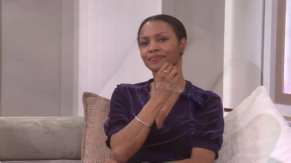 A woman in a deep purple velvet top smiles gently, her fingers touching her chin. The QVC Extra logo is visible on a screen behind her, suggesting she's on a broadcast. A woman in a deep purple velvet top smiles gently, her fingers touching her chin. The QVC Extra logo is visible on a screen behind her, suggesting she's on a broadcast.