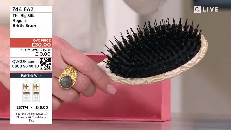 A hand, adorned with a large grey stone ring, holds up a black bristle hairbrush. The brush, with its cream-coloured handle, is angled over a bright pink box.