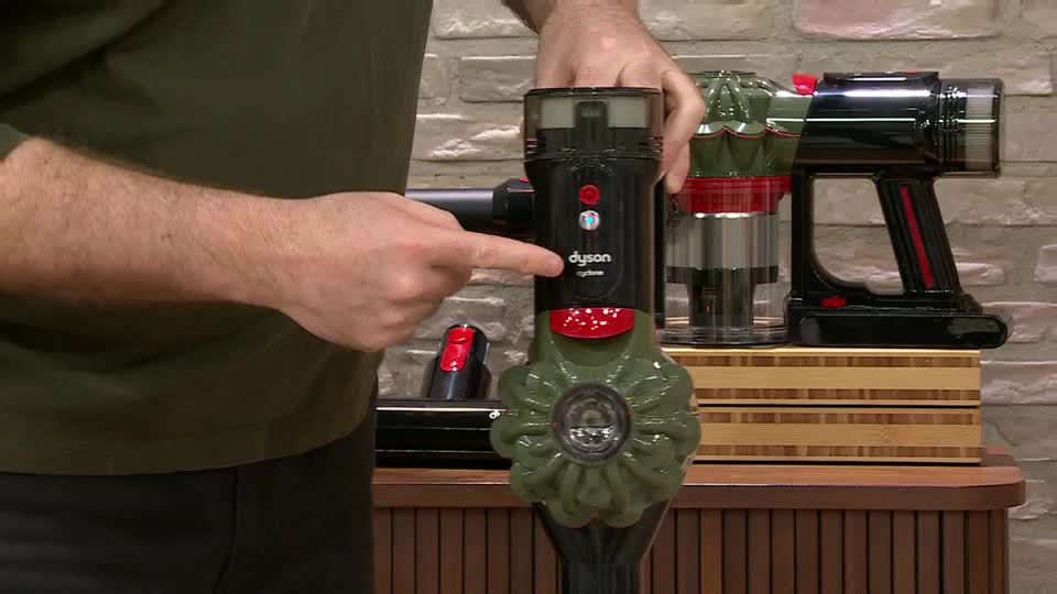 A man's hand points to the "dyson" logo on a green and black vacuum cleaner. Another Dyson vacuum, with a clear canister, sits to its right.