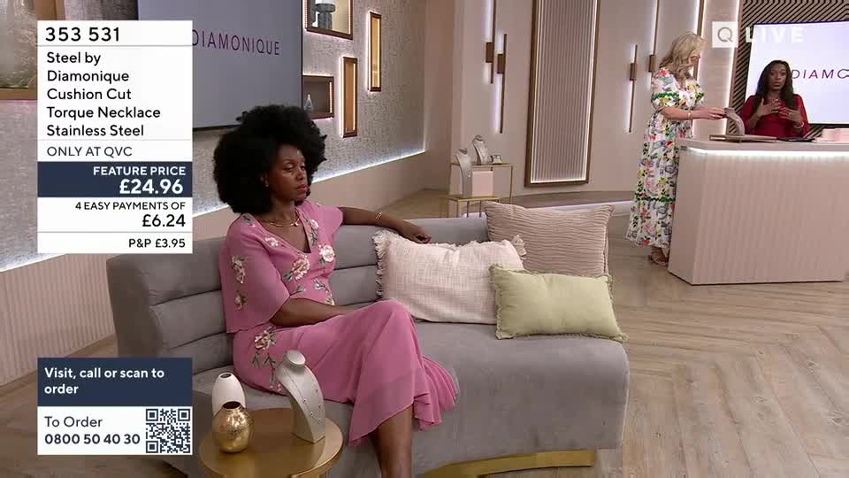 A woman in a pink floral dress is sitting on a sofa, looking off to the side. Across the room, another woman in a white floral dress stands behind a counter, gesturing as she speaks.