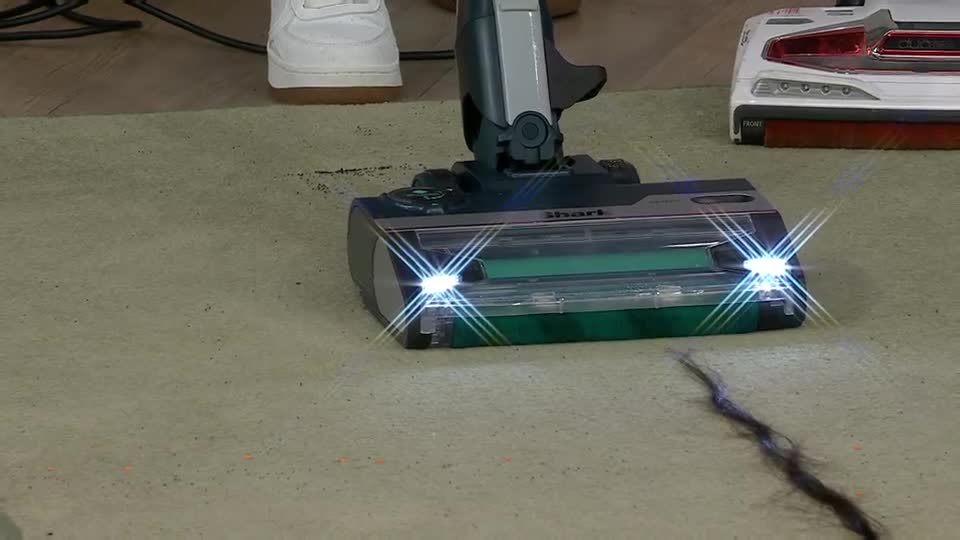 The vacuum head, with its bright LED lights illuminating the carpet, glides forward. A long, dark strand of debris is being drawn into the machine.