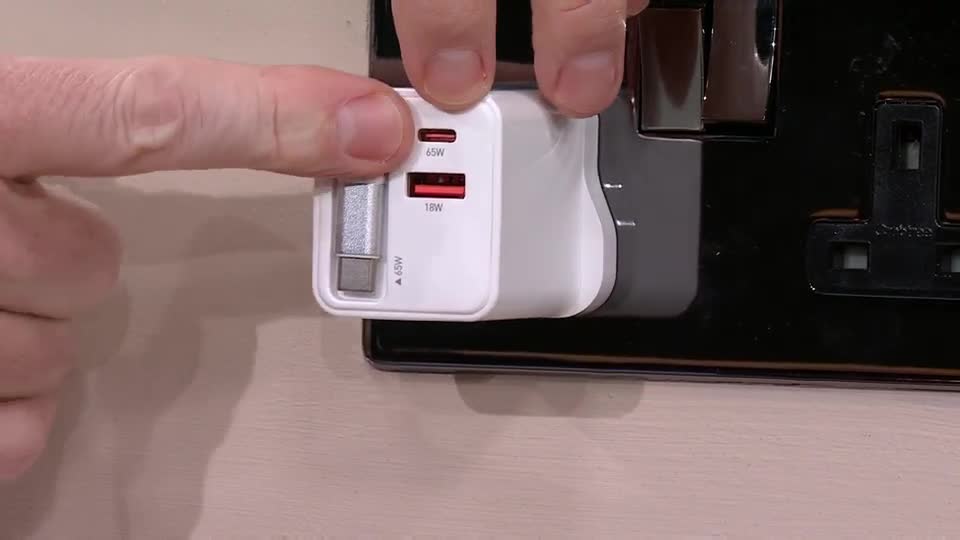 Fingers push a white charging brick into a black UK electrical socket. The brick has two USB-C ports, one red and one grey, and a silver prongs.