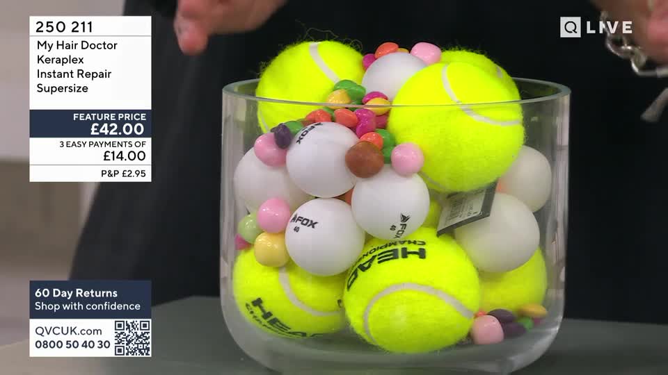 A clear glass vase overflows with bright yellow tennis balls, interspersed with white ping pong balls and a scattering of colourful sweets. A hand reaches in from the right, its fingers just visible near the top of the arrangement. A clear glass vase overflows with bright yellow tennis balls, interspersed with white ping pong balls and a scattering of colourful sweets. A hand reaches in from the right, its fingers just visible near the top of the arrangement.