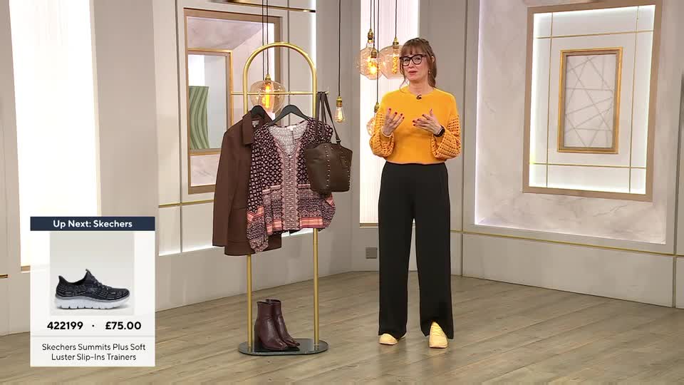 A presenter stands in a studio, gesturing with her hands while a rack of clothing and a display of shoes sit nearby. A graphic overlay shows a pair of Skechers trainers and their price.