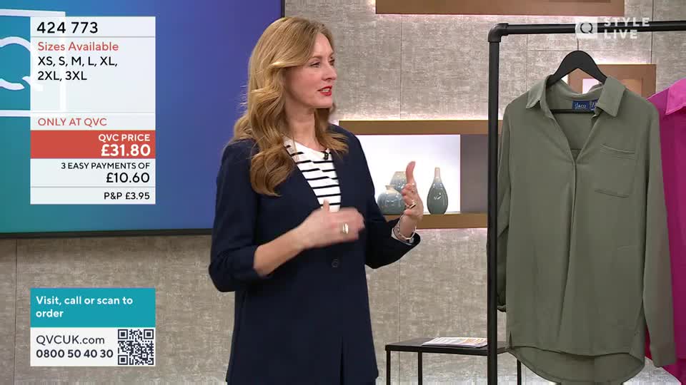 A woman in a navy blazer and striped top gestures animatedly as she presents a khaki-coloured shirt hanging on a rack. The QVC price of £31.80 is displayed on a screen behind her.