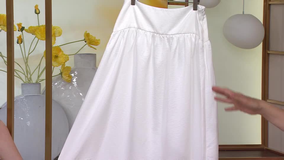 A hand reaches out, brushing against the flowing white fabric of a skirt. Yellow poppies stand in a vase behind it, a cheerful splash of colour against the muted backdrop.