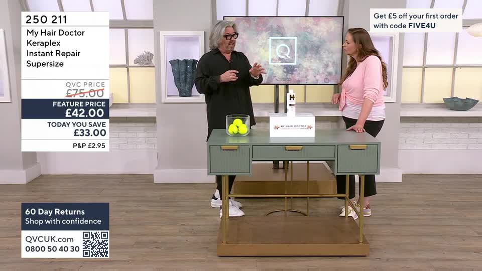 A man in a black tunic gestures animatedly while a woman in a pink cardigan stands nearby, both on a QVC set. A display for "My Hair Doctor Keraplex Instant Repair Supersize" sits on a green desk between them.