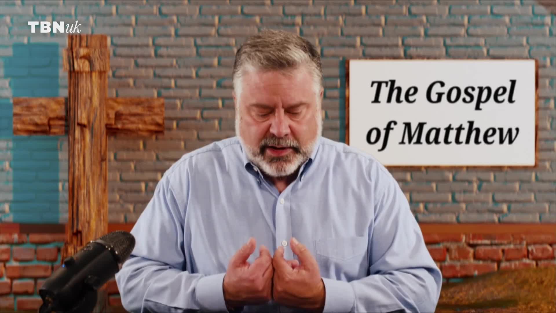 A man with a grey beard stands with his hands clasped, eyes closed, before a wooden cross. A sign behind him reads "The Gospel of Matthew" on TBN UK.