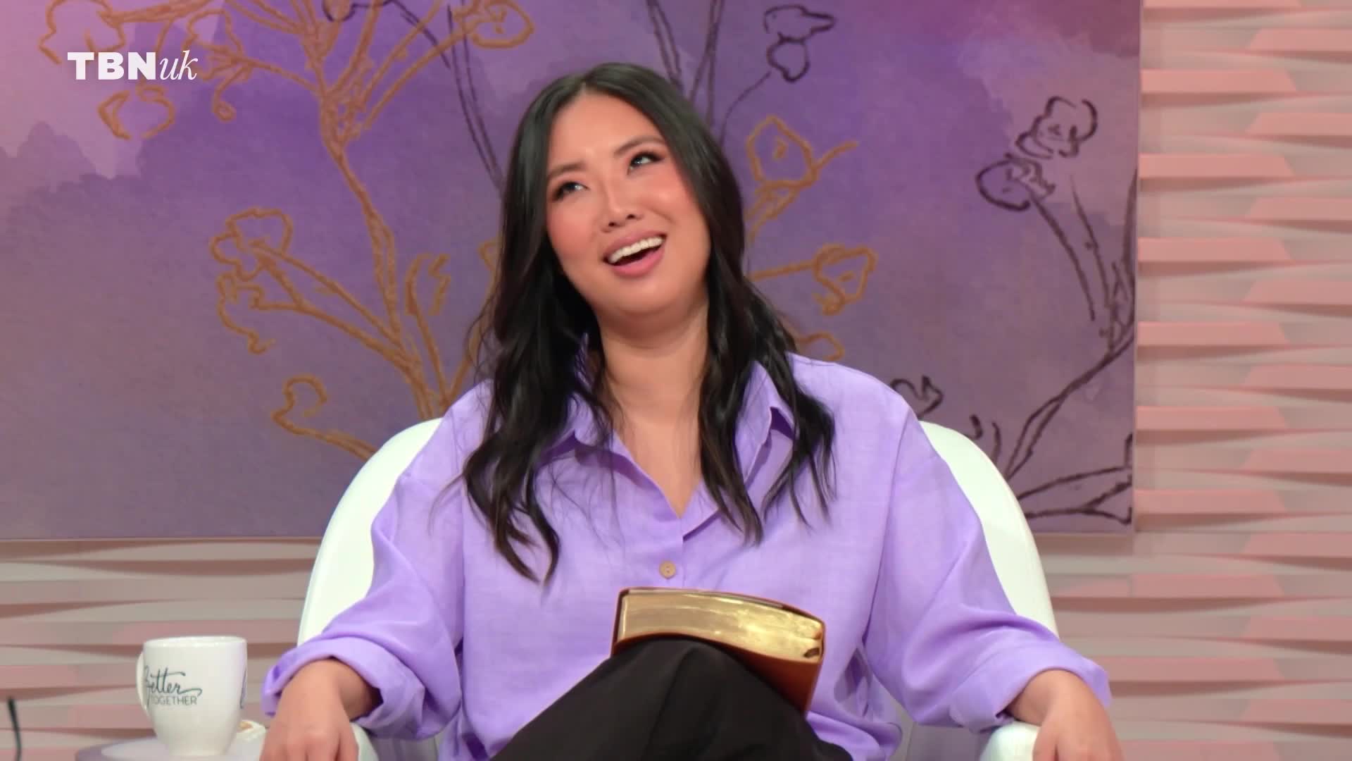 A woman in a lilac shirt is speaking animatedly, a gold-edged book resting on her lap. The TBN UK logo is visible in the upper left corner.