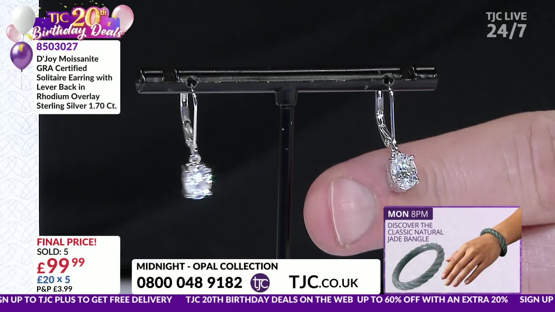 A hand holds a dangling silver earring with a clear stone. Another identical earring hangs from a display stand beside it.