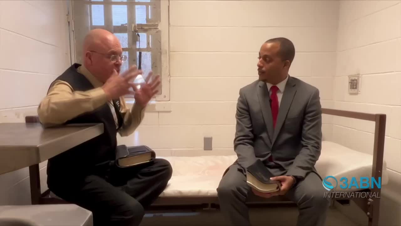 A man in a vest gestures emphatically with his hands, speaking to another man in a suit. Both men sit in what appears to be a prison cell, each holding a Bible.
