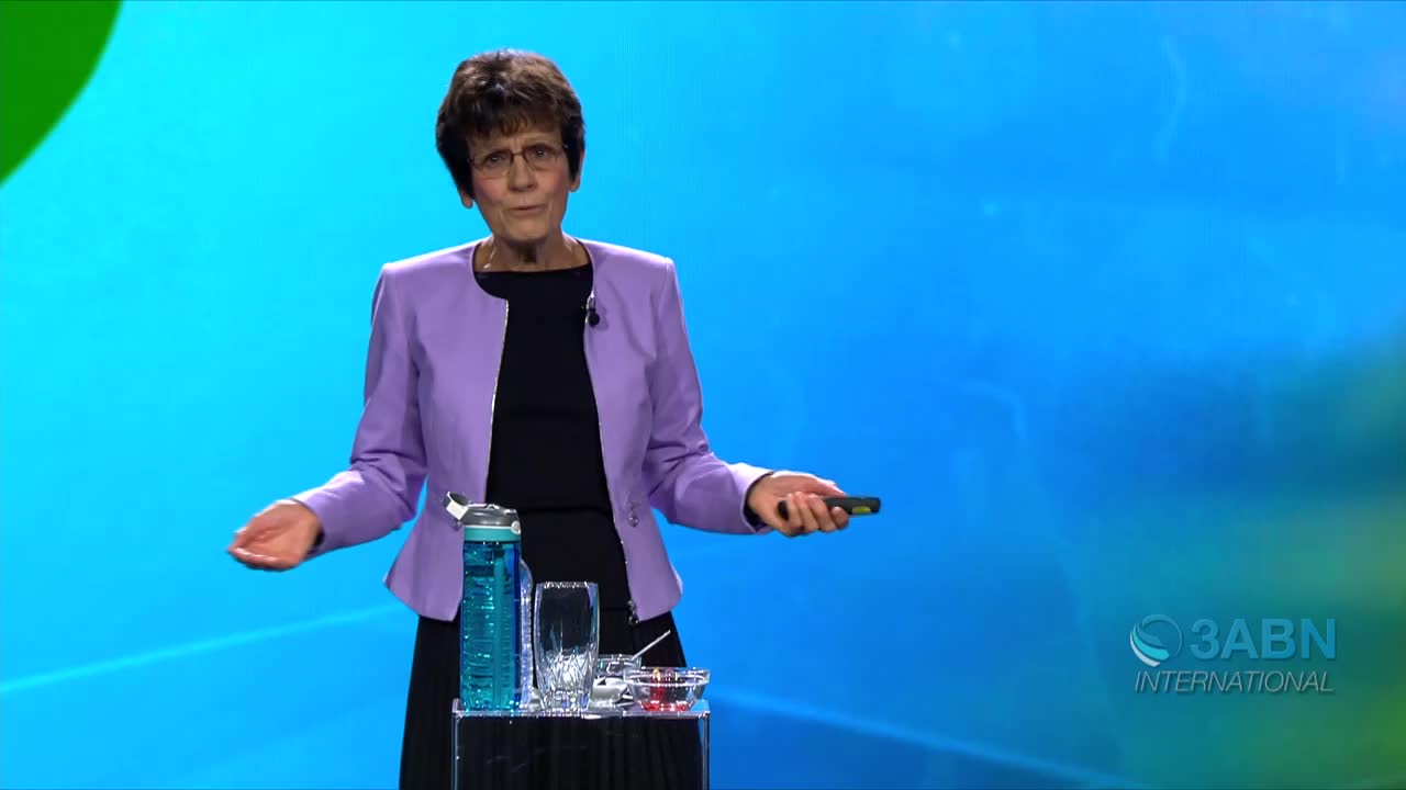 A woman in a purple jacket stands on a stage, gesturing with her hands. Behind her, the 3ABN International logo is visible against a blue backdrop.
