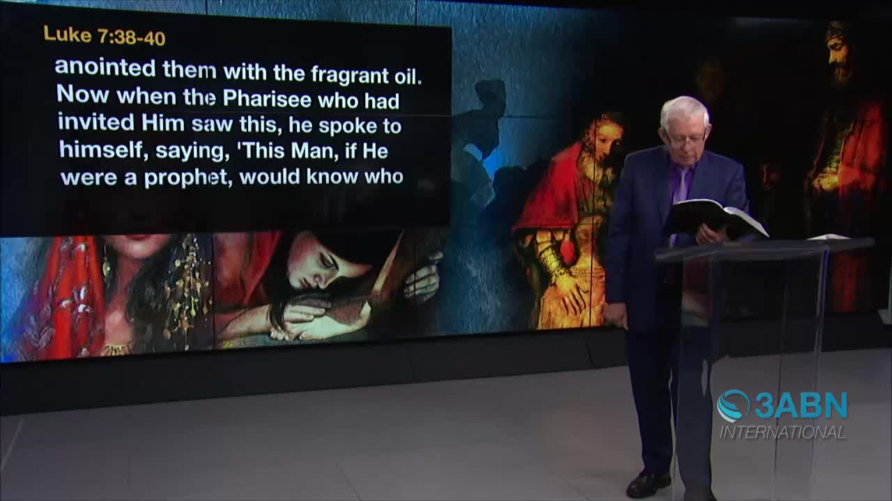 A man in a dark suit stands at a lectern, reading from a book. Behind him, a screen displays text and images, and the 3ABN International logo is visible.
