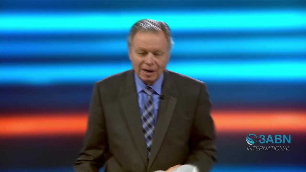 A man in a suit, with a blue and gray tie, is speaking into a microphone. Behind him, a backdrop of blue, orange, and white stripes is visible, and the 3ABN International logo is in the bottom right corner.
