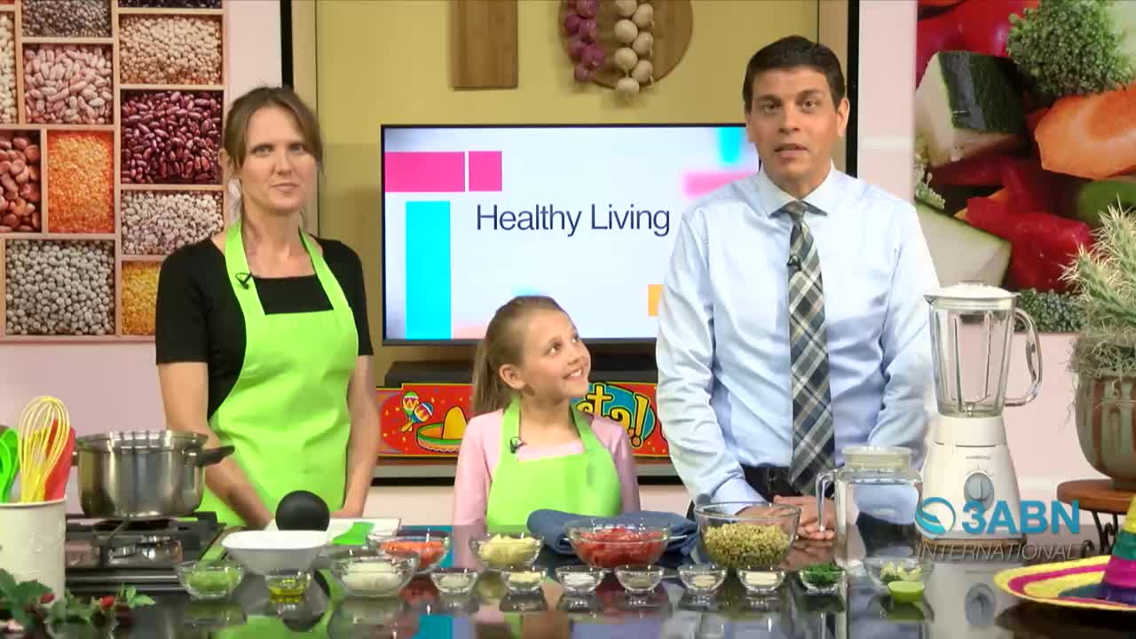 A woman, a girl, and a man stand behind a table of ingredients, all wearing aprons. The 3ABN International "Healthy Living" set has a blender and a pot on the stove.
