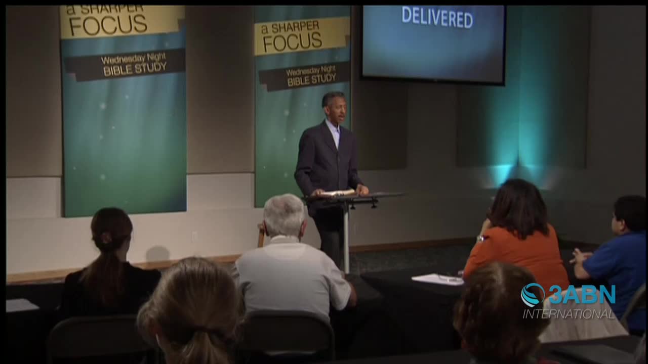 A man in a suit stands behind a lectern, speaking to an audience. Behind him, a screen displays the word "DELIVERED," and the 3ABN International logo is visible in the corner.
