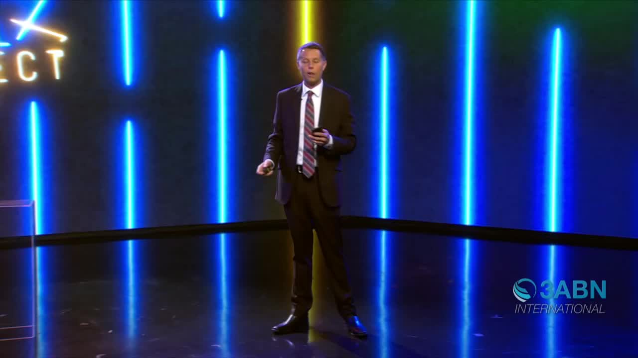 A man in a suit stands center stage, holding a phone and gesturing. Behind him, vertical neon blue lights illuminate the backdrop for a 3ABN International broadcast.
