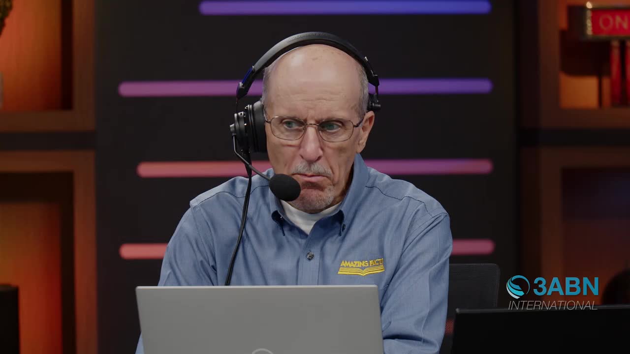 A man with headphones on leans forward, his brow furrowed in concentration. He's wearing a blue shirt with a yellow logo, and a laptop sits open in front of him.