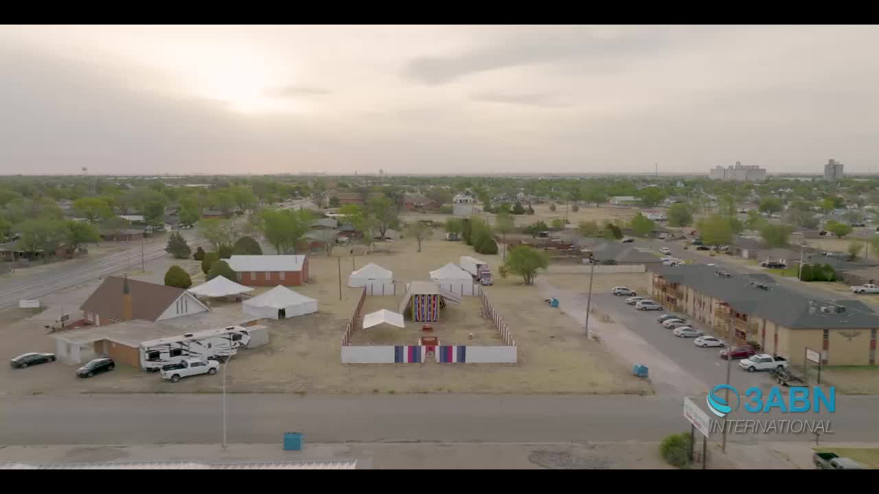 A temporary encampment of white tents stands in a dusty lot, surrounded by a low fence. Several vehicles, including RVs and cars, are parked nearby.