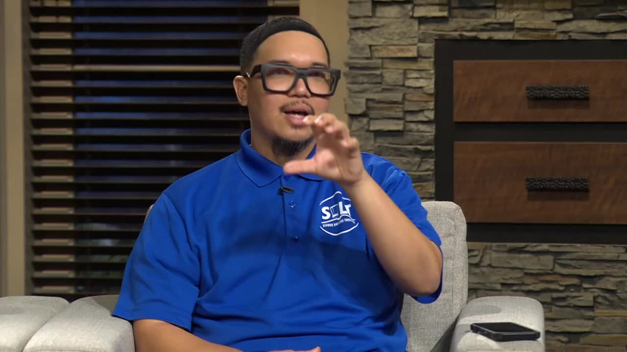 A man in a bright blue polo shirt with the 3ABN International logo gestures with his hands. He's seated on a couch, speaking animatedly.