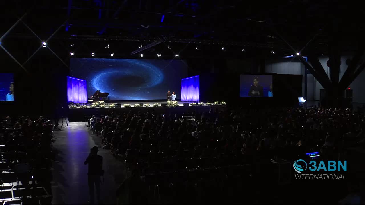 Two speakers stand on a brightly lit stage in front of a swirling blue galaxy projection. A large audience fills the darkened hall, their faces illuminated by the stage lights.