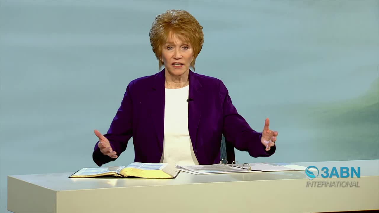 A woman in a purple jacket speaks with her hands gesturing. A Bible lies open on the table in front of her.
