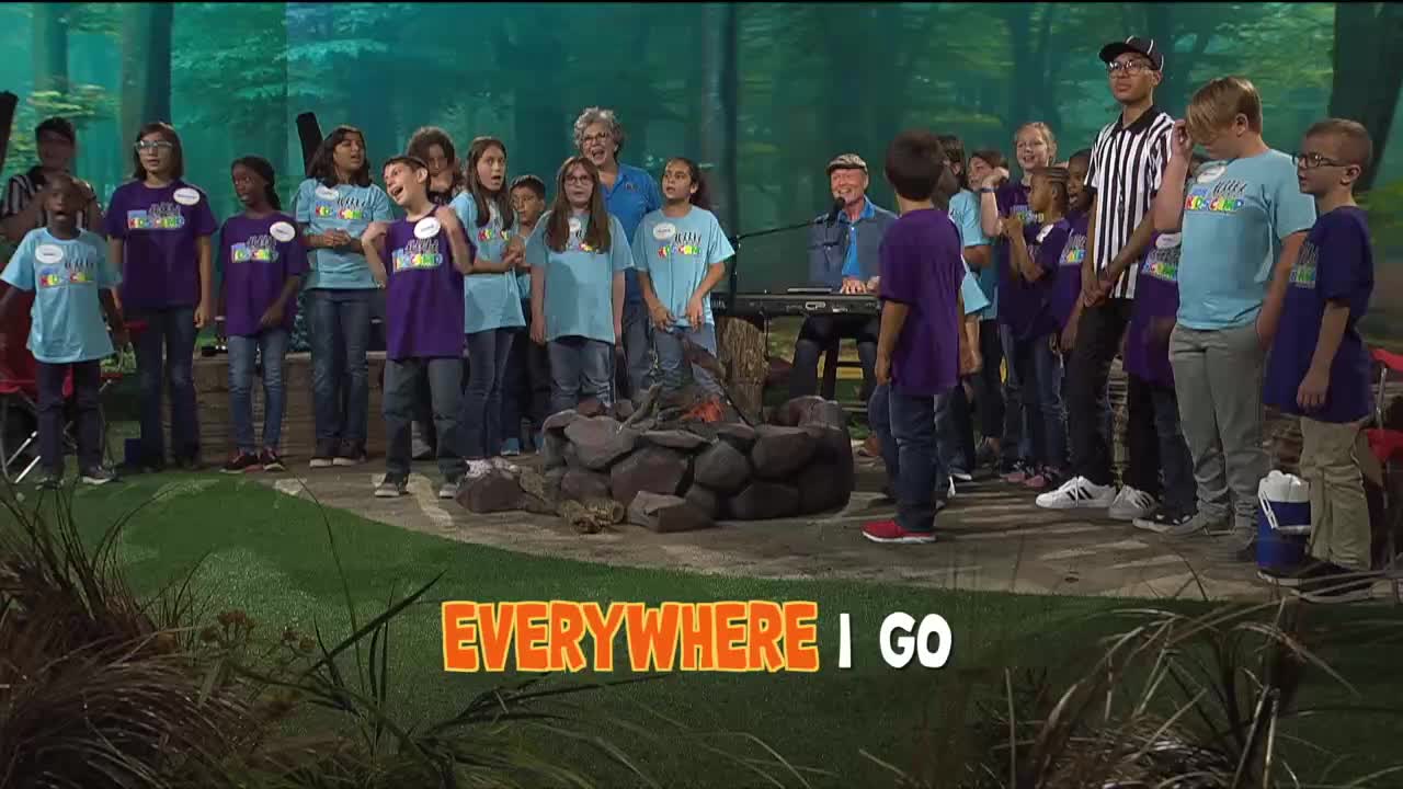 A group of children in matching shirts sing, some with their hands raised, in front of a faux campfire. A man plays a keyboard, and a referee stands to the side, watching the performance.

