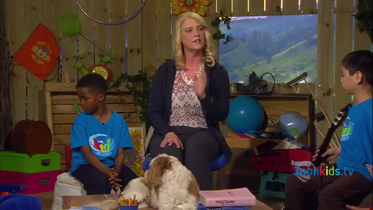 The woman in the center gestures with her hands as she speaks. Two children, wearing blue t-shirts, sit on either side of her, one with a guitar.

