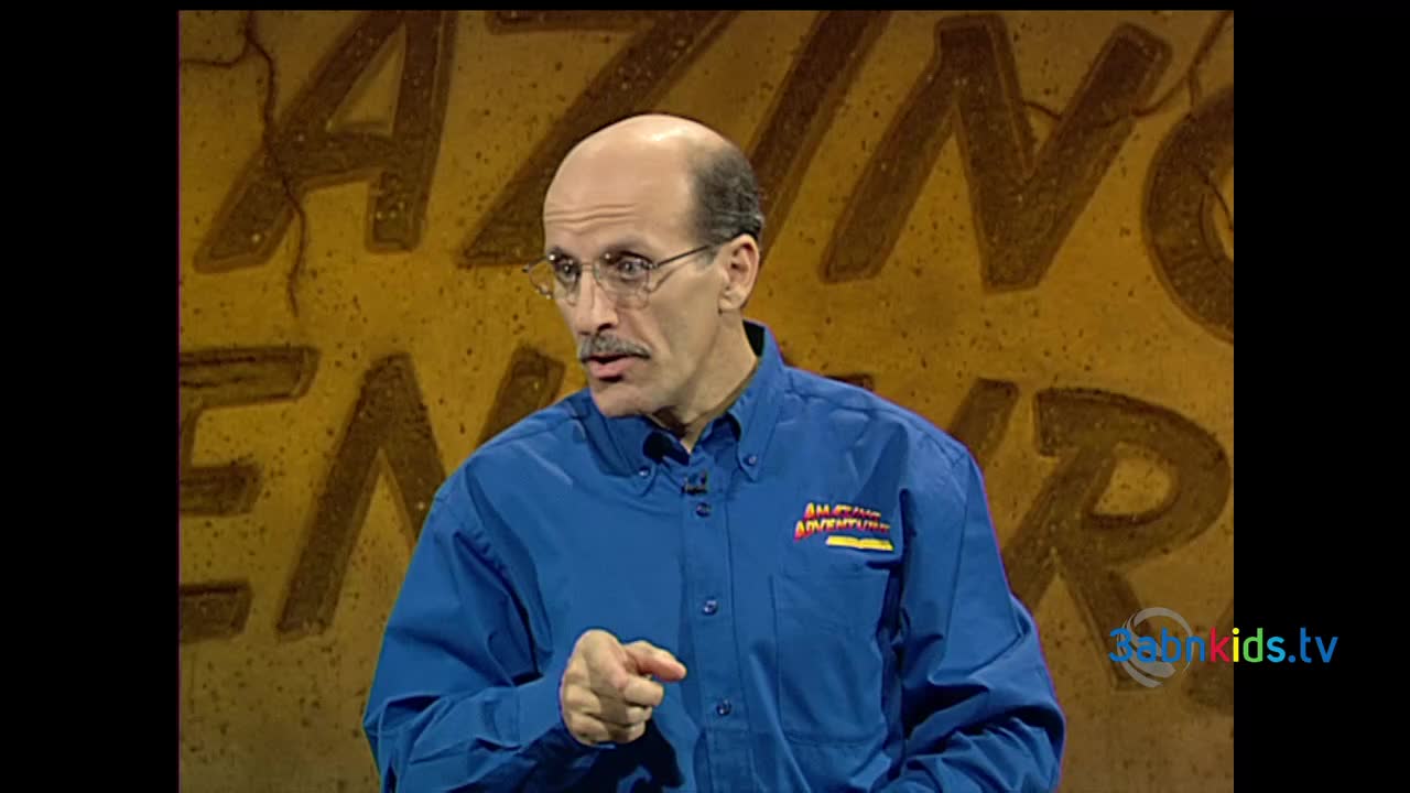 The man in the blue shirt, wearing glasses, is pointing directly at something off-camera. Behind him, the wall is painted with large, stylized letters.
