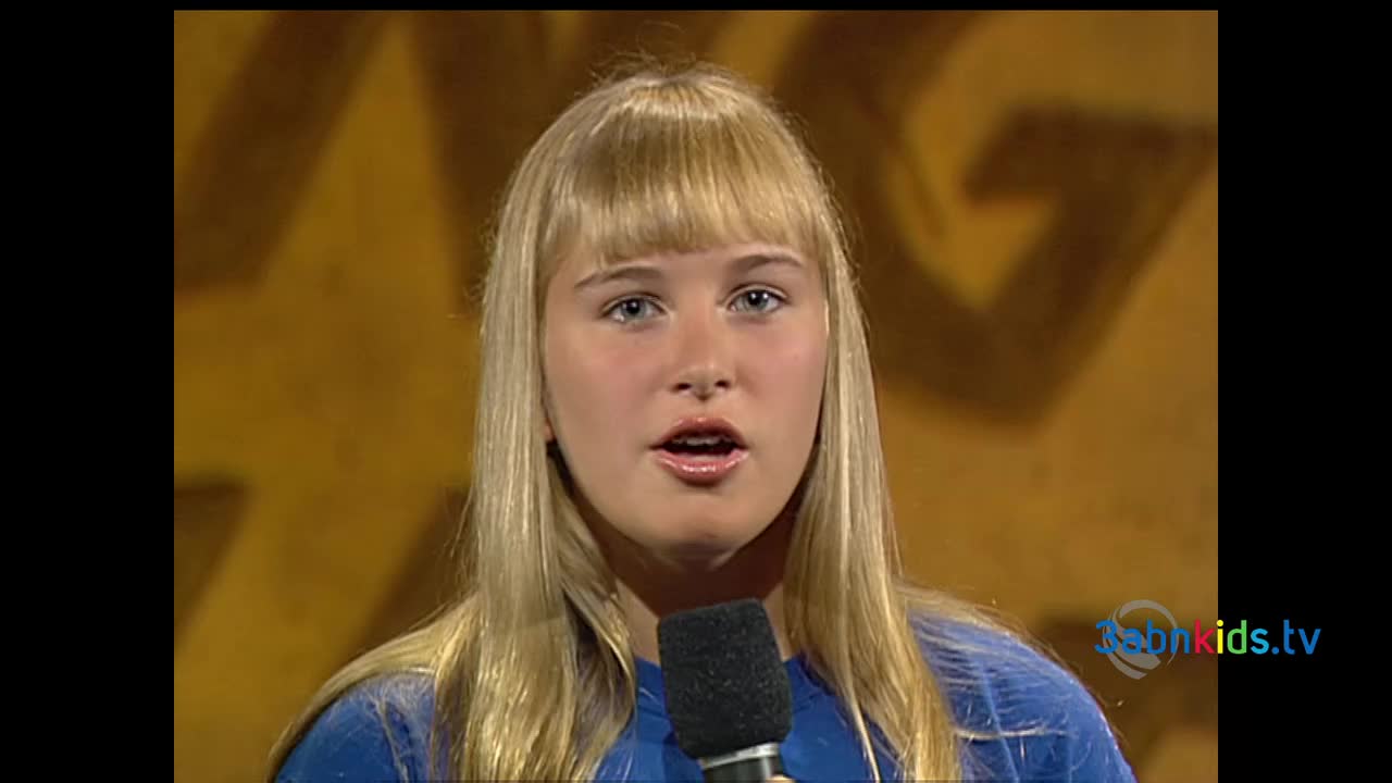 A young woman with blonde hair and blue eyes is speaking into a microphone. Behind her, a gold-colored background features a large "G" shape, and the logo for 3ABN Kids is visible in the corner.
