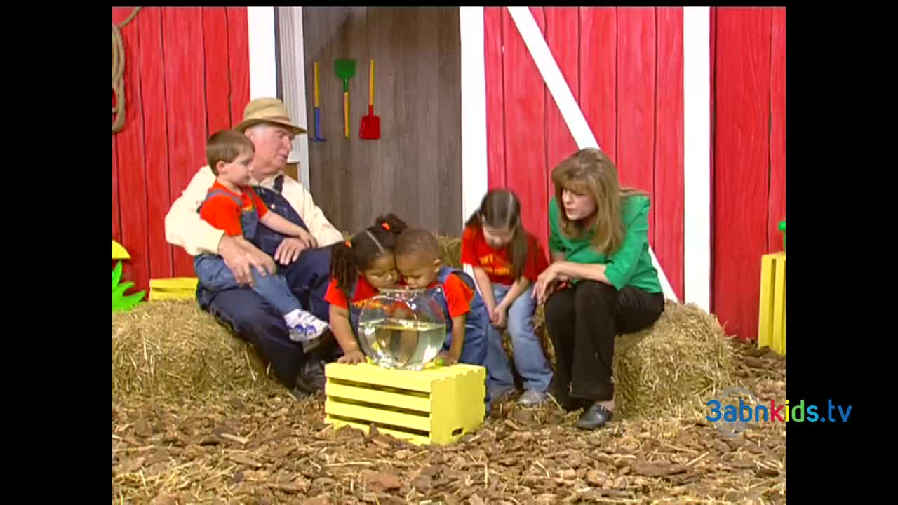 A group of children in red shirts and denim overalls are gathered around a fishbowl on a yellow crate. A man in a straw hat and a woman in a green shirt are sitting nearby, watching them.
