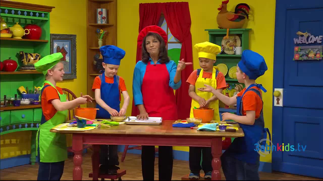 A woman in a red apron and chef's hat gestures towards a table where children in colorful aprons and hats are mixing ingredients. The scene is set in a brightly decorated kitchen, part of a 3ABN Kids program.
