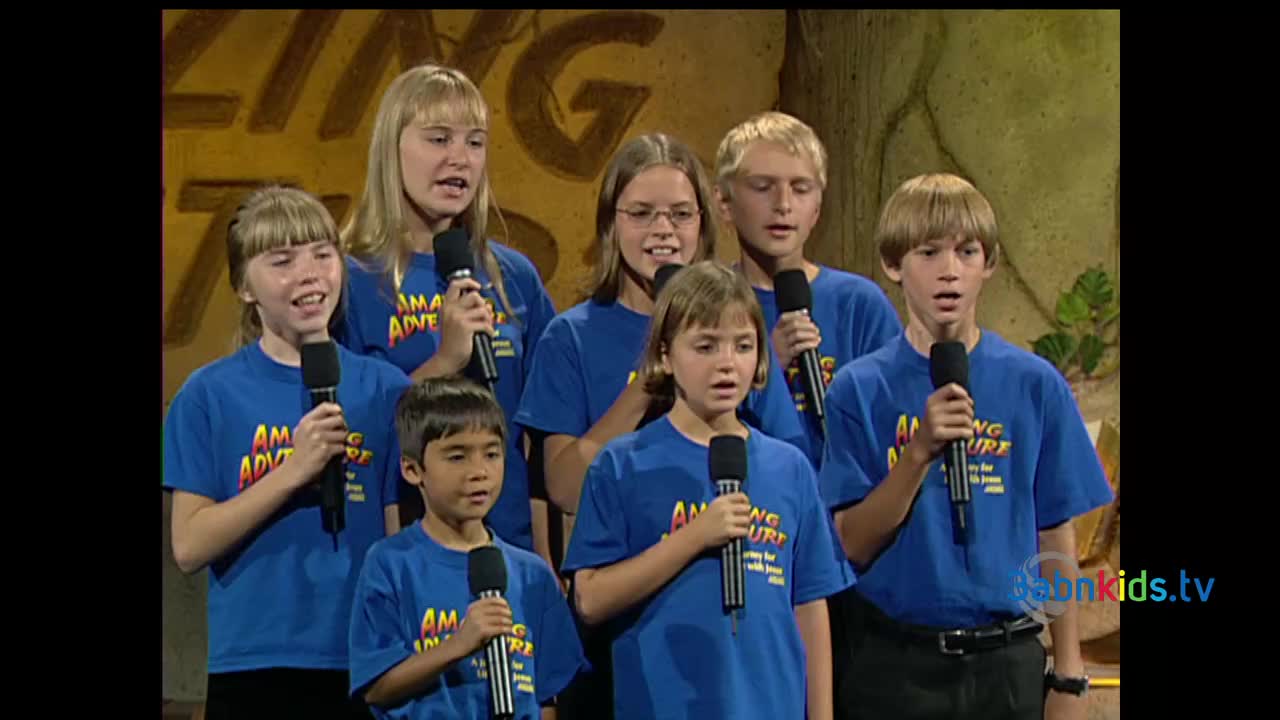 A group of children, all wearing blue t-shirts, are singing into microphones on a stage. The backdrop is a painted wall with the words "AMAZING ADVENTURE" in large letters.
