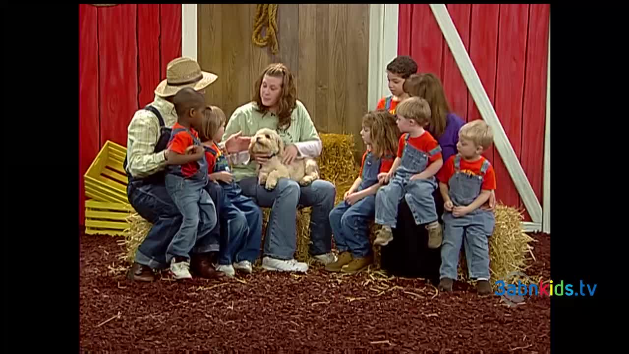 A group of children and adults sit on hay bales in front of a barn backdrop. A woman holds a small, fluffy dog while talking, and the kids are looking on with interest.
