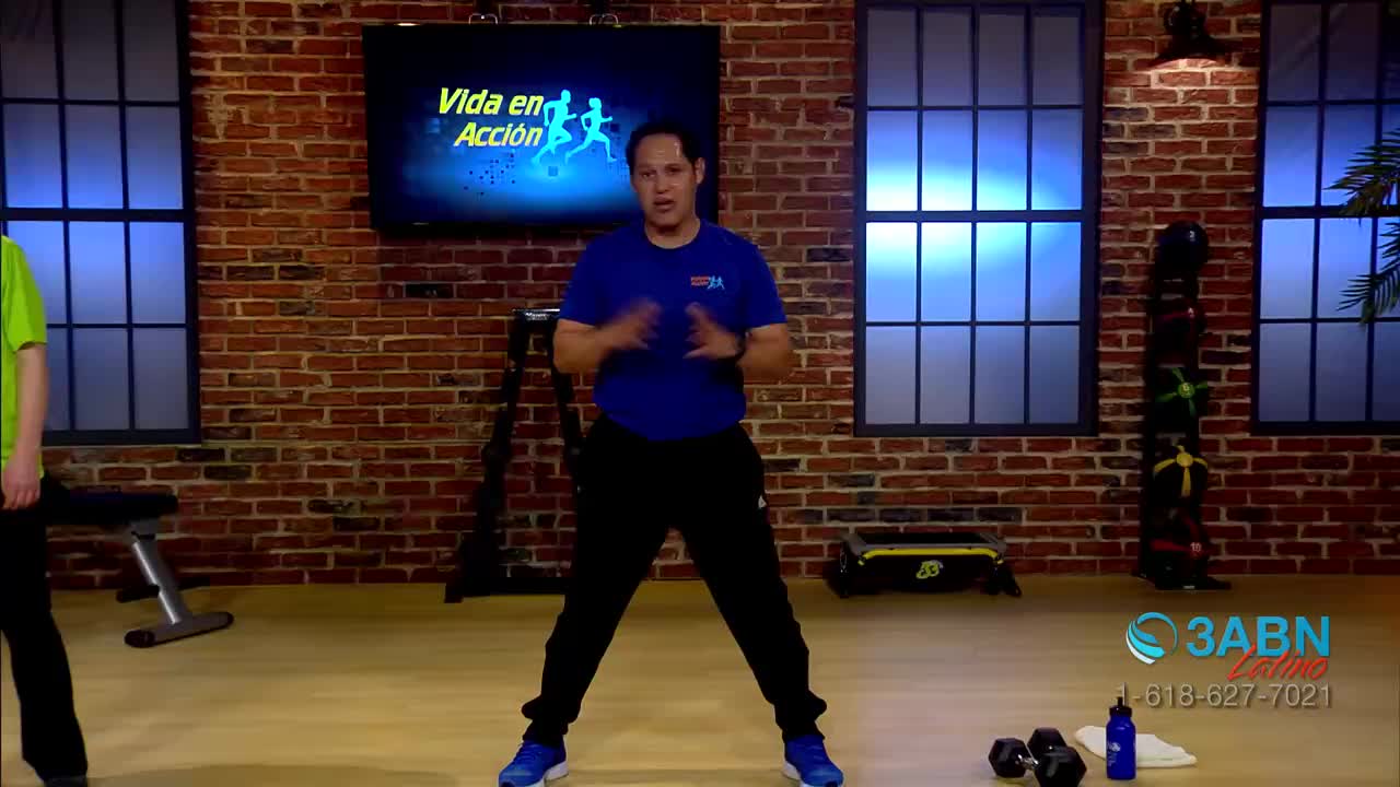 A man in a blue shirt and black pants is demonstrating an exercise, his arms moving in front of him. Behind him, a screen displays the words "Vida en Acción," and the 3ABN Latino logo is visible to the right.
