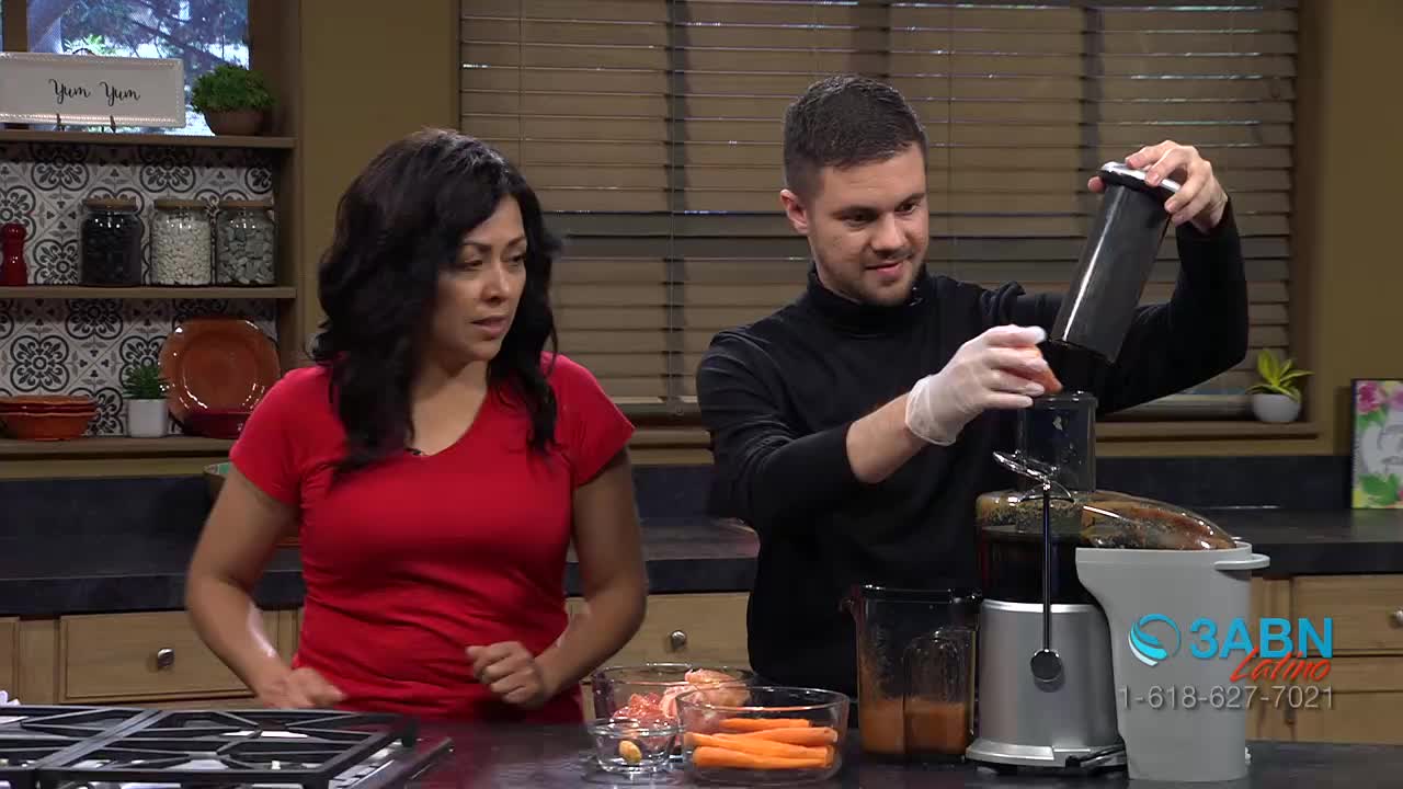 A man in a black turtleneck is carefully placing a part of the juicer back in place, while a woman in a red shirt watches. The scene is set in a kitchen, with the 3ABN Latino logo visible in the corner.
