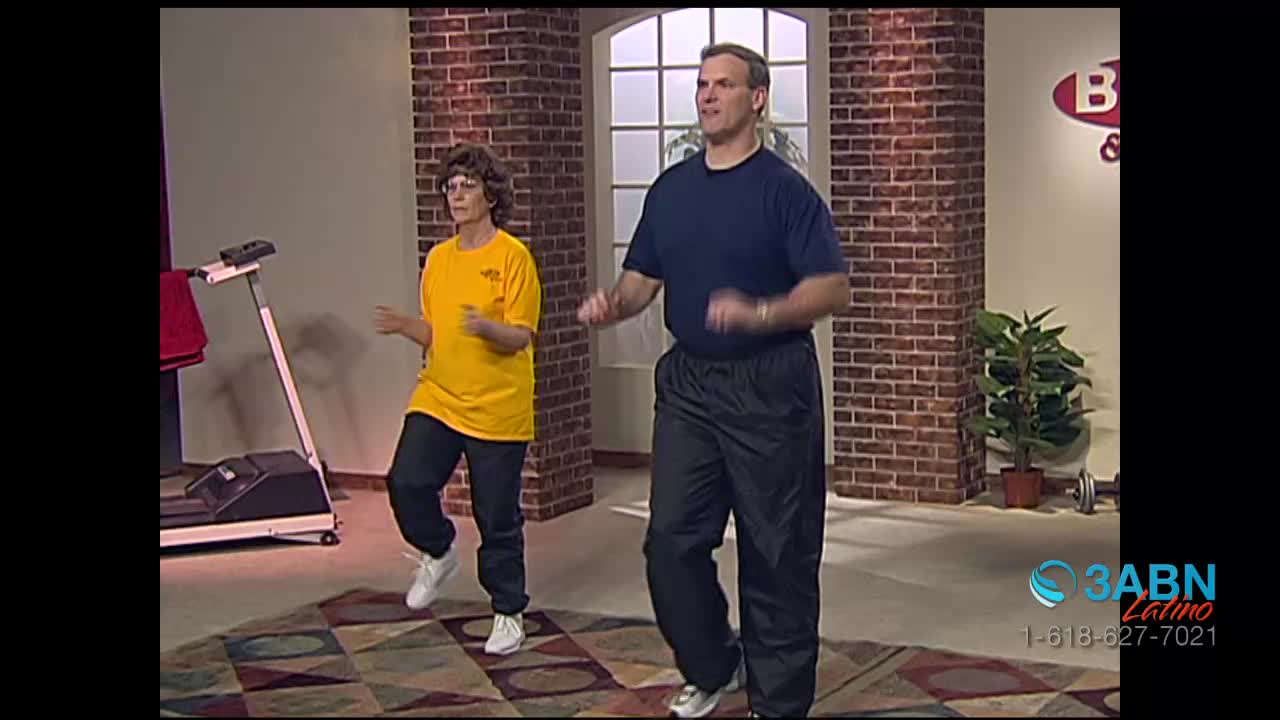 Two people, a man and a woman, are jogging in place on a patterned rug. The woman wears a yellow shirt and the man wears a dark blue one; behind them is a treadmill and a brick wall.
